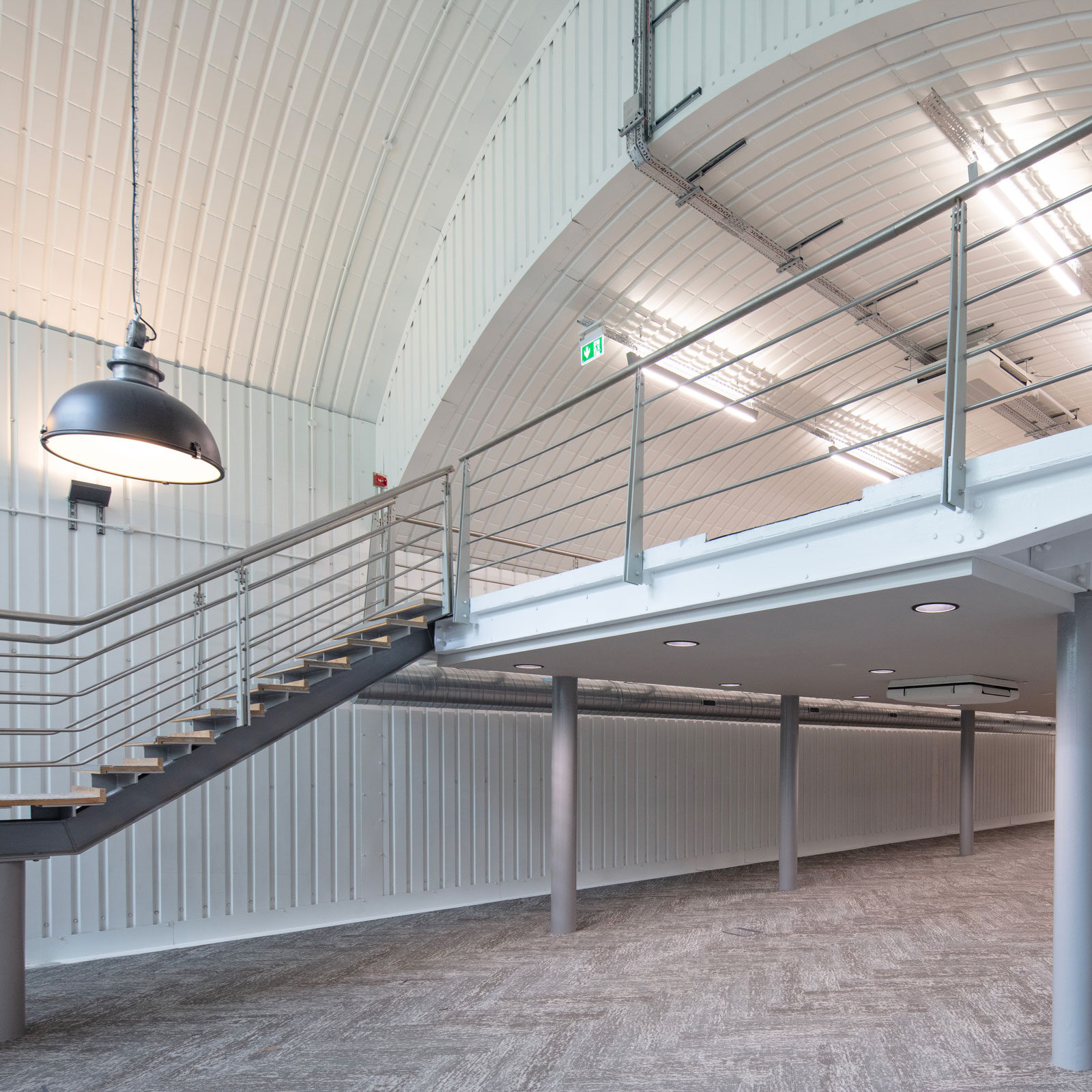 Modern industrial interior with a metal staircase leading to a mezzanine. Curved ceiling, carpeted floor, and hanging pendant lights create an open, airy feel.